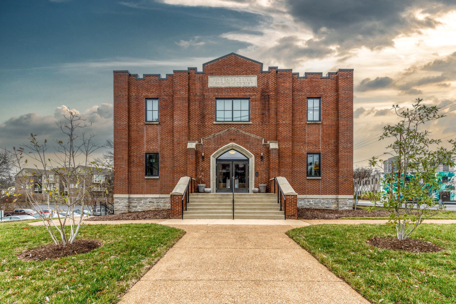 The brick facade of Riverside Station, a former 1937 church, its stepped parapet and pointed-arch entrance rising behind a walk and a flight of stone steps at dusk.