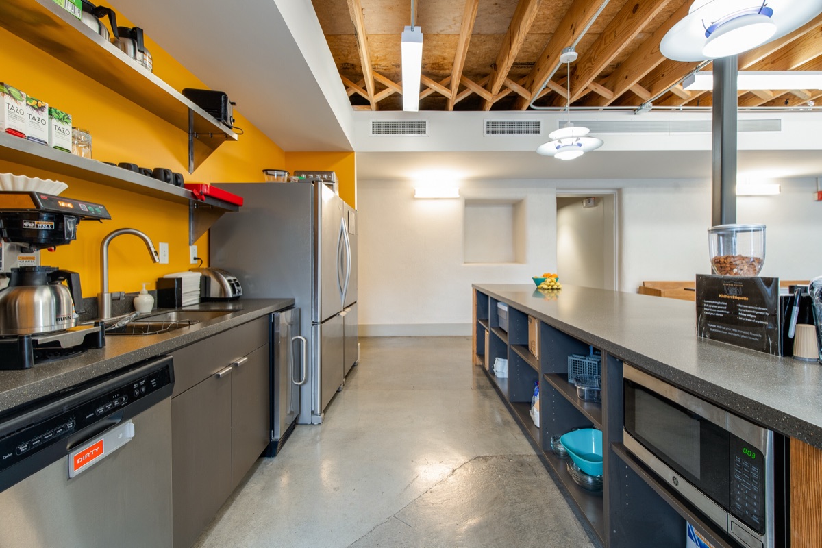 The shared kitchen, with coffee makers and open shelving against a mustard wall, and a long counter of storage cubbies opposite.