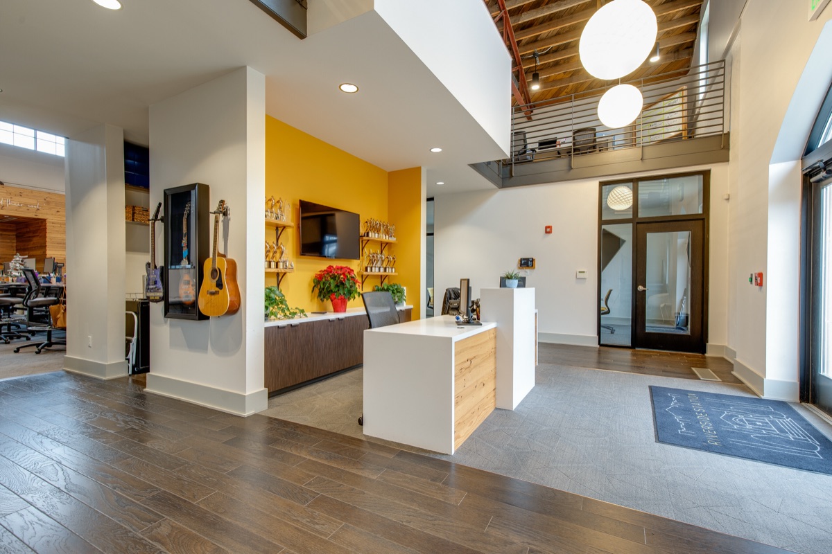 The reception desk under the mezzanine, with guitars mounted on the wall, a mustard accent wall behind a wall-mounted screen, and the arched front doors to the right.