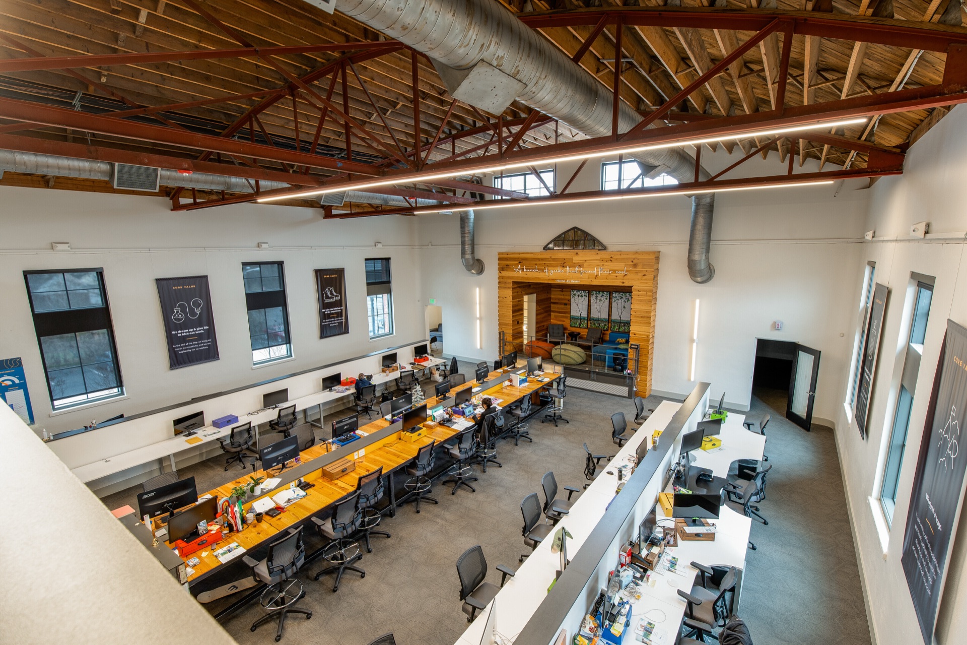 The main level seen from the mezzanine: a long reclaimed-wood work table running the length of the room beneath the exposed ceiling and its red steel trusses.